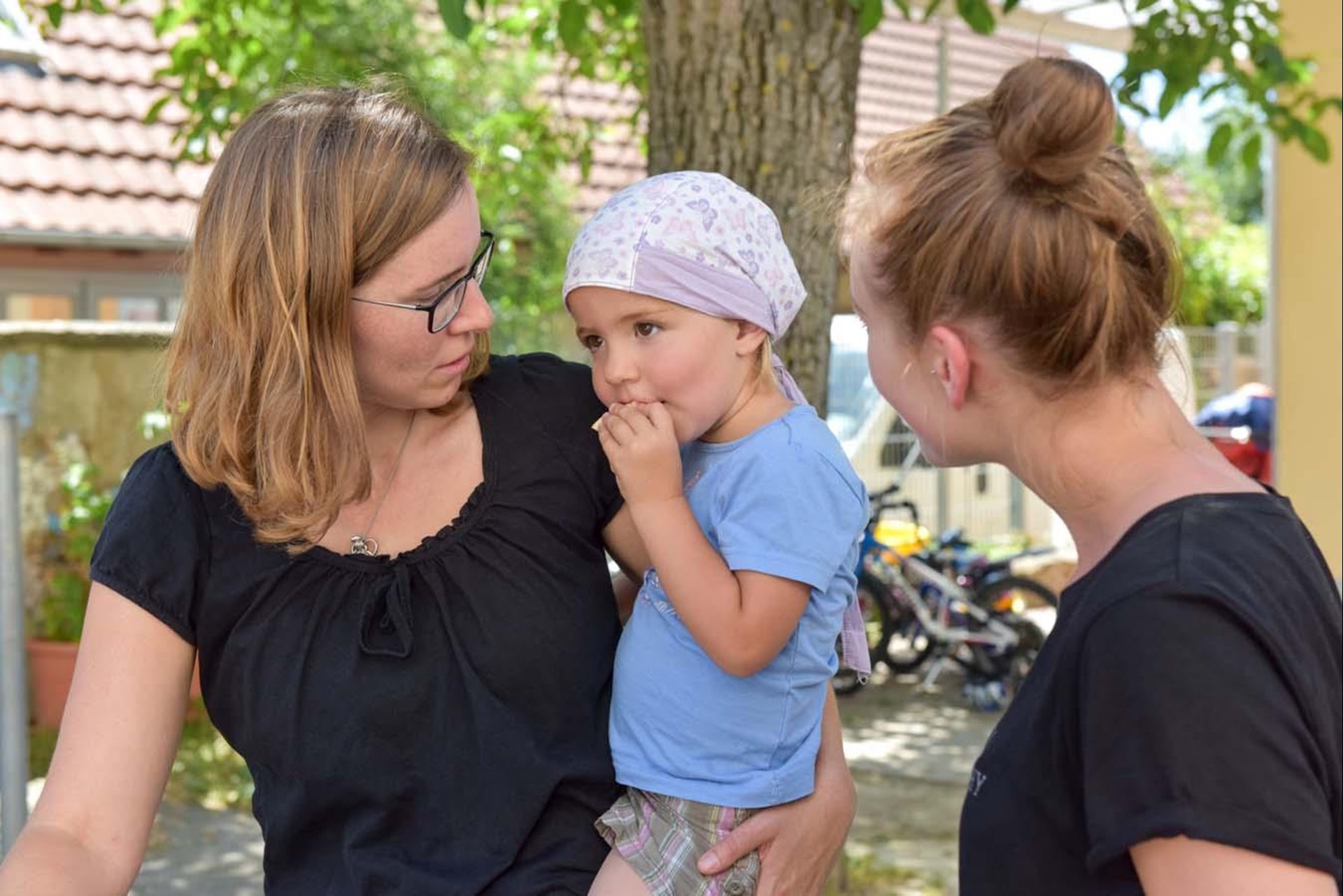 Mutter mit Tochter auf dem Arm und Erzieherin stehen vor einer Kindertagesstätte