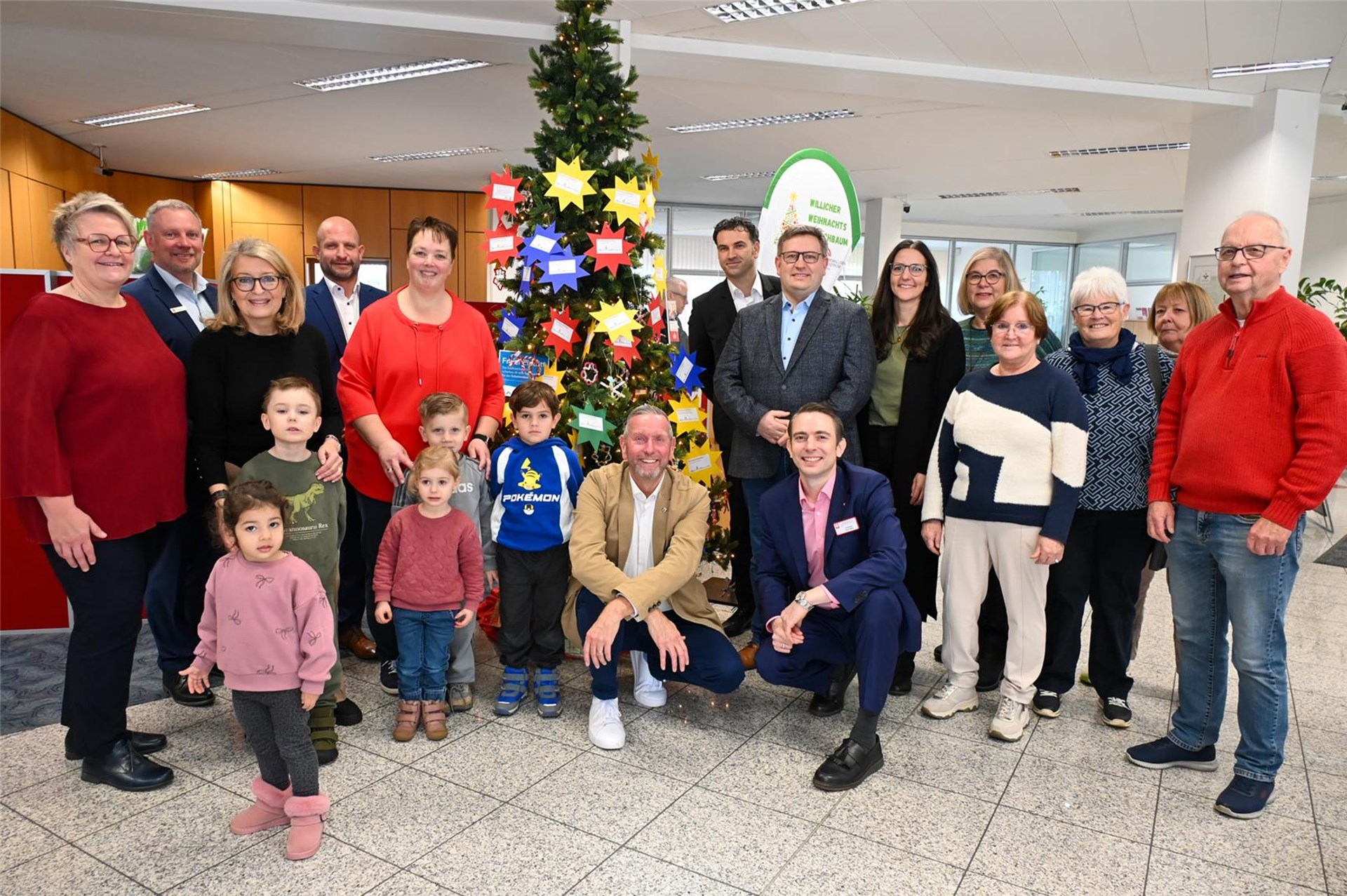 Gruppenfoto von Erwachsenen und Kindern vor einem geschmückten Weihnachtsbaum in einem Innenraum mit Büroumgebung. Der Baum ist mit bunten Sternen und Wunschzetteln behängt. Die Gruppe wirkt fröhlich und festlich, einige Personen knien oder hocken vorne, während andere stehen. Im Hintergrund sind Büromöbel, Pflanzen und eine weihnachtlich dekorierte Tafel sichtbar.